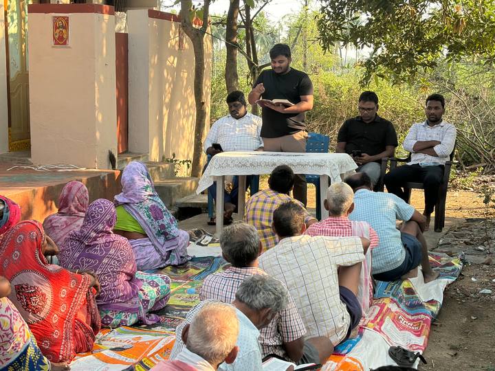 People seated on mats in a yard while a young man reads aloud at a table
